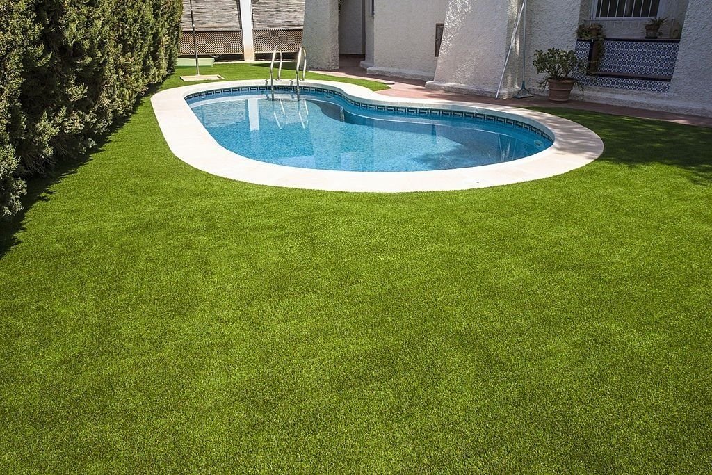 A backyard with a small, kidney-shaped swimming pool surrounded by white pavement. The well-manicured green grass from Mesa Artificial Grass complements the serene space beautifully. Trees on the left provide shade, while the exterior of a white house with a window and a potted plant adds charm on the right. Free estimates available for Artificial Grass Landscaping projects.