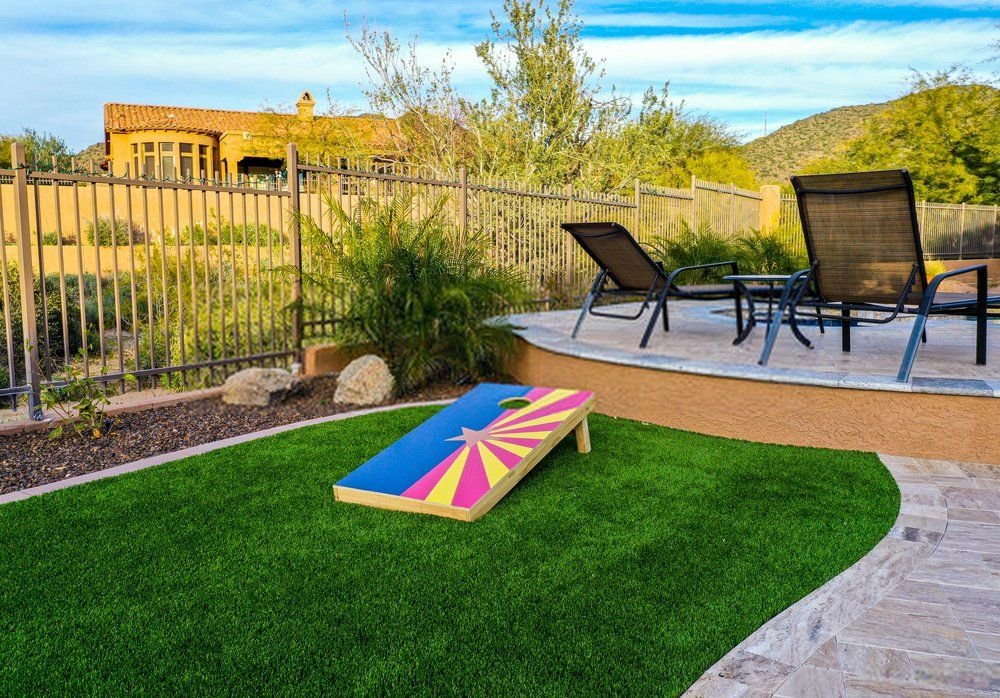 A backyard scene with two lounge chairs on a patio near a fence, overlooking a desert landscape. In the foreground, a cornhole board with a vibrant design sits on pet-friendly artificial grass for easy clean-up. Lush green plants and a distant house under a blue sky complete the view.