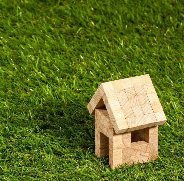 A small wooden toy house is placed on a lush green lawn made of artificial grass. The house is simple and constructed from interlocking wooden pieces. The scene suggests a natural, outdoor setting with vibrant greenery and a hand-crafted, rustic toy ideal for backyard putting greens.