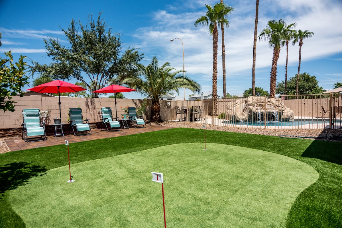 A backyard scene featuring a green putting practice area with two flag-topped holes in the foreground. In the background, furnished by the top-rated artificial grass landscape company in Mesa, AZ, there are lounge chairs with red umbrellas, palm trees, and a swimming pool with a rock waterfall feature under a clear blue sky.