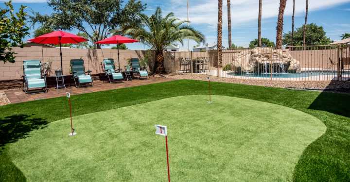 A backyard in Gilbert AZ features a mini-golf putting green with flags, surrounded by synthetic grass solutions. To the left, lounge chairs with red umbrellas rest on a patio, and in the background, palm trees and a pool with a waterfall complete this idyllic desert oasis.