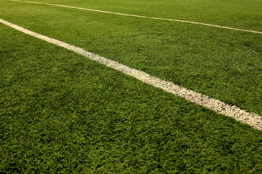 A close-up view of green grass on a sports field resembles an artificial grass installation, with two parallel white lines running diagonally across the image. The turf appears well-maintained and vibrant under natural light.