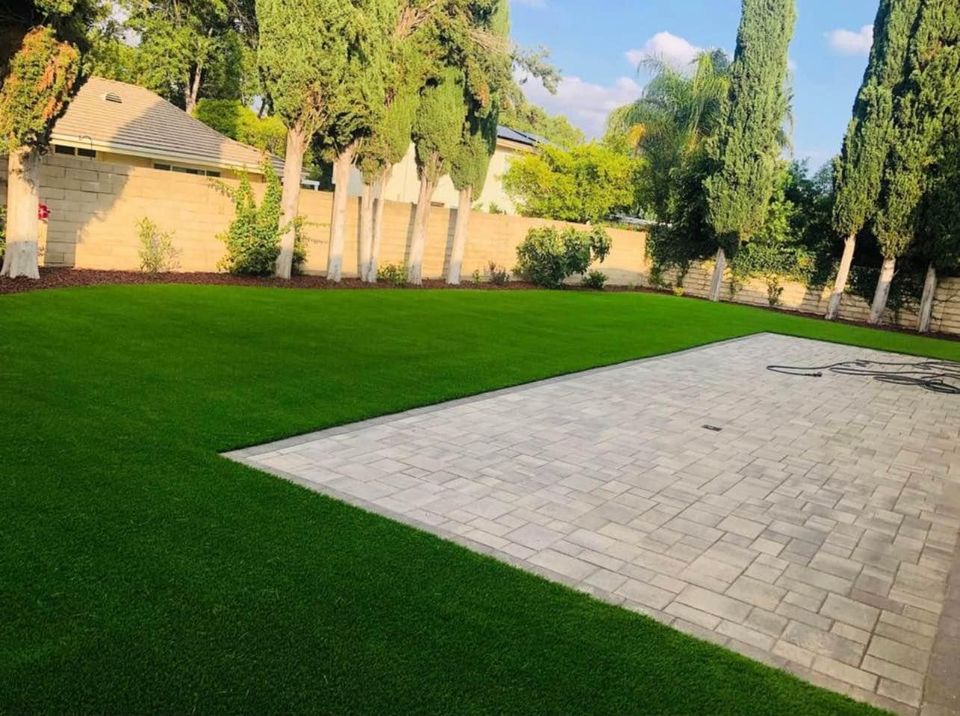 View of a backyard with neatly maintained putting greens and a paved patio area. Tall trees and a stone wall line the perimeter, creating a peaceful setting. The sun casts gentle shadows, highlighting the vibrant colors of the lawn's lush greenery and garden.