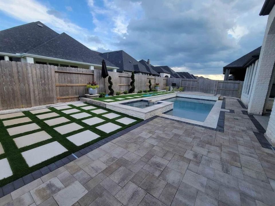 A backyard patio in Queen Creek is transformed into a low-maintenance oasis, featuring a modern swimming pool with an integrated hot tub. The area boasts stone-tile surfaces, geometric artificial grass patterns, and potted plants. Encircled by a wooden fence, it rests against suburban rooftops under cloudy skies.