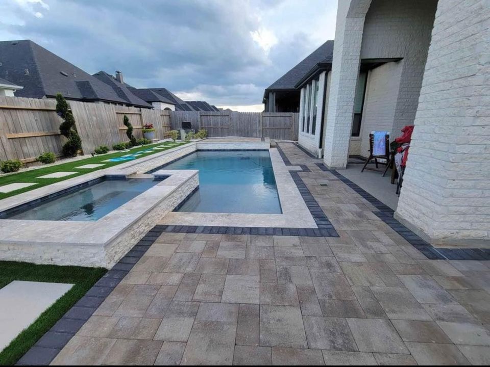 A backyard in Apache Junction features a rectangular swimming pool and spa surrounded by stone tiles. Enclosed by a wooden fence, the area boasts artificial turf installation for a low-maintenance lawn. Manicured shrubs enhance the scene as a cloudy sky casts soft light overhead.