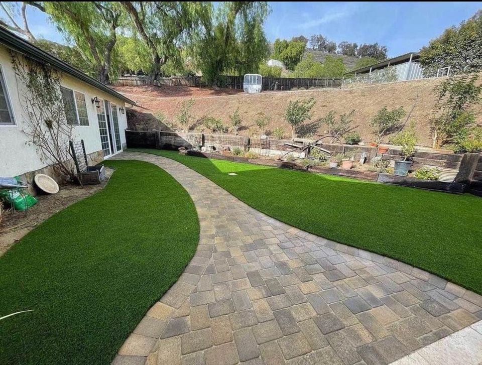 A backyard features a stone pathway winding through sections of well-manicured pet turf. The area is bordered by a raised garden bed with various plants. A house with a slanted roof is to the left, and trees line the hillside in the background.