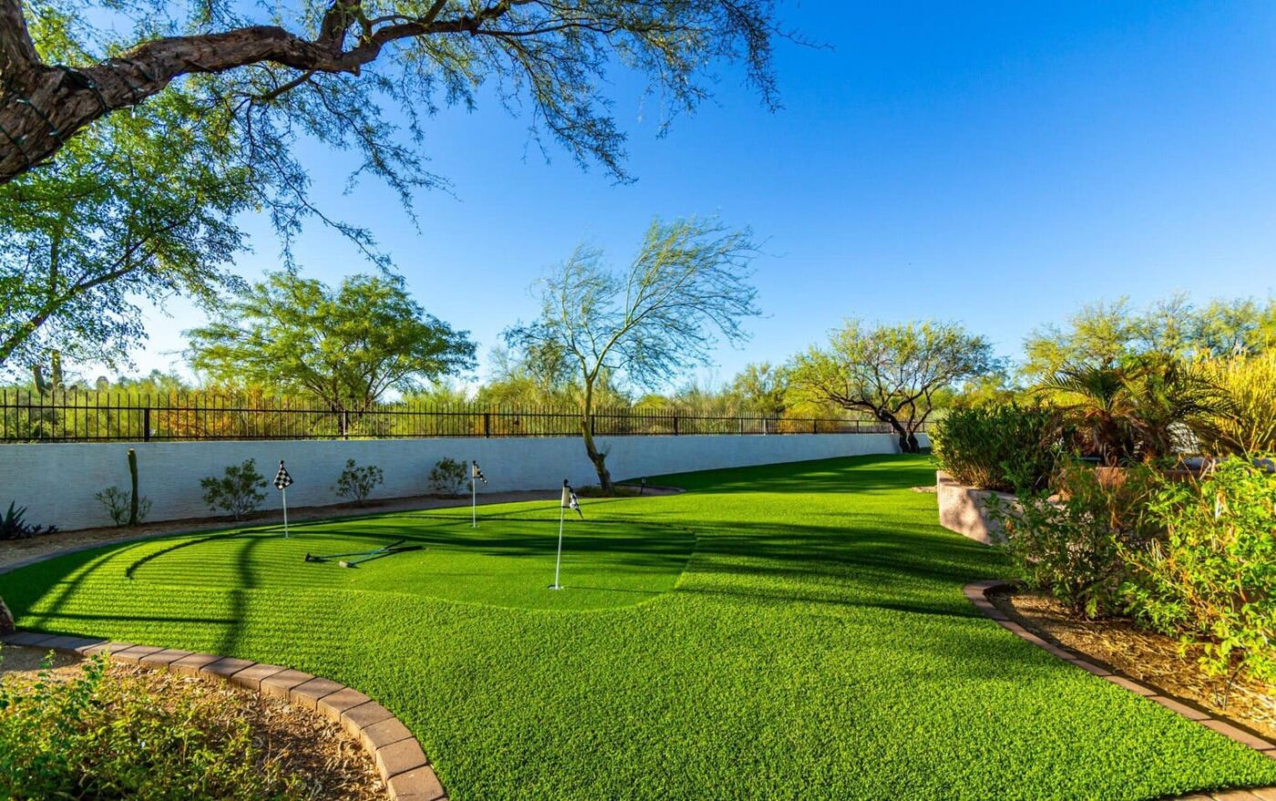 A lush backyard in Apache Junction boasts a premium installation featuring a putting green surrounded by trees and a brick border. The artificial turf green includes several golf holes with flags, set against a blue sky and enclosed by a white fence in the sunny, landscaped area.