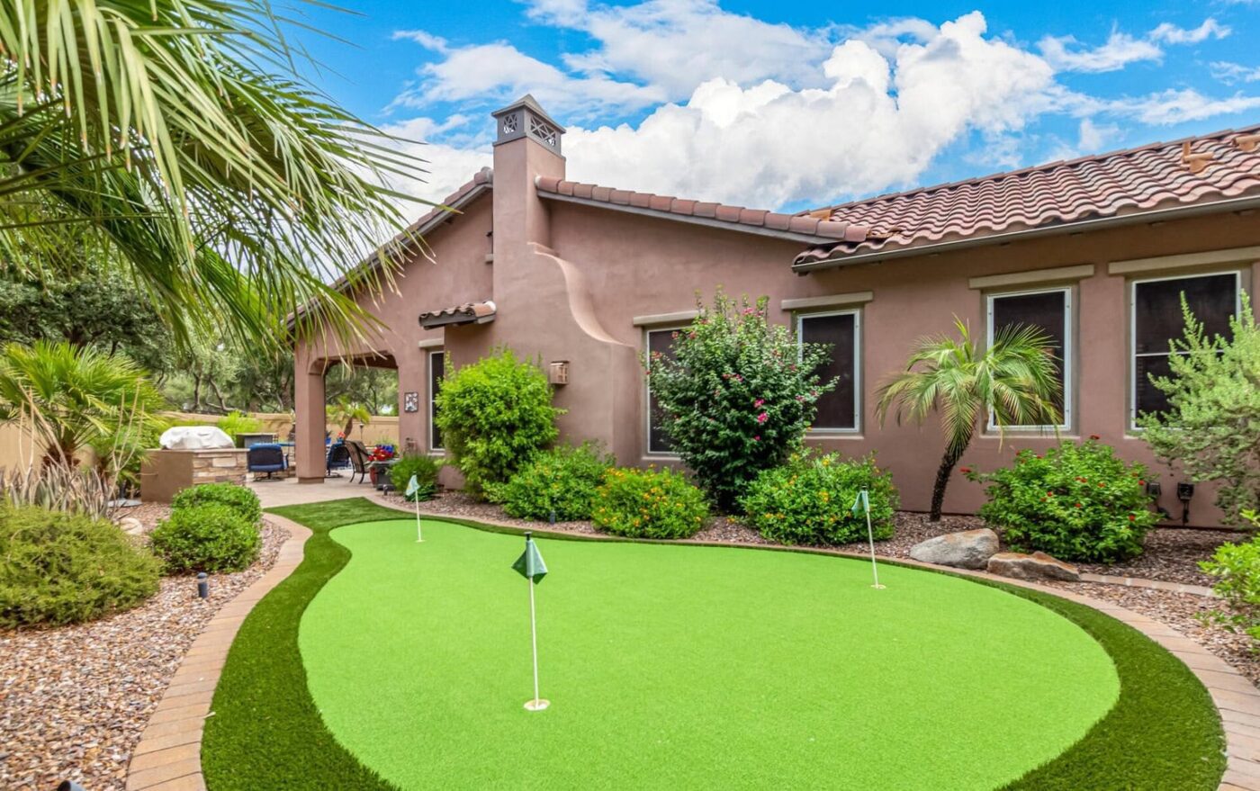 A backyard scene features a small putting green with artificial grass installation, surrounded by desert landscaping. The house boasts a stucco exterior with red roof tiles, framed by various plants and palm trees. The sky is bright with scattered clouds, perfect for enjoying this serene oasis.